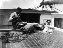 Baby on shipboard with a seaman who is repairing a sail